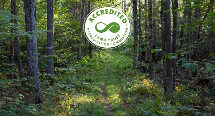 Land Trust Accreditation Commission seal over a hiking trail in a northern deciduous forest