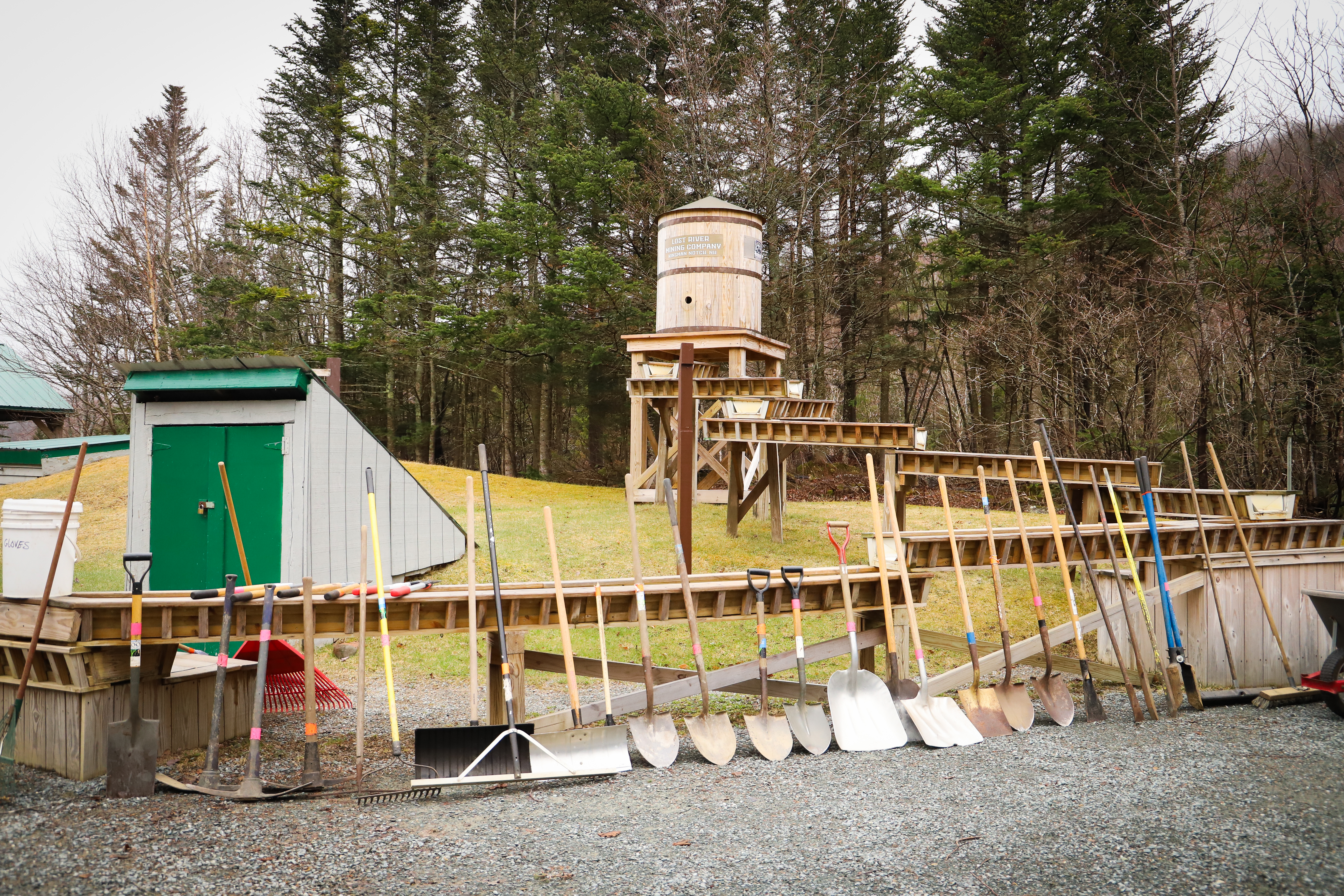 A line of shovels, rakes and other garden tools lean against a wooden fence