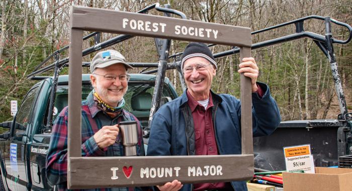 Two men smile from behind a wooden frame that says "Forest Society I <3 Mt Major"