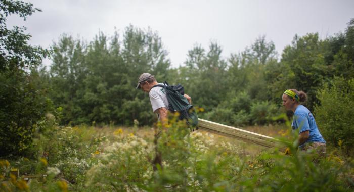 Man and woman carry large wooden post through field