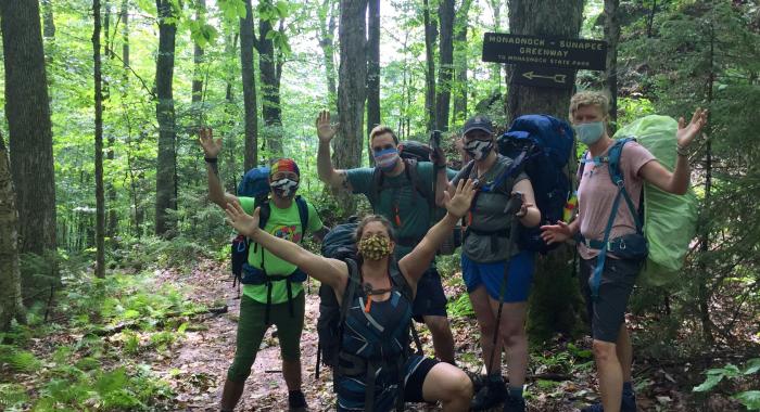 A group of masked hikers poses in front of a sign on the Monadnock-Sunapee Greenway trail.