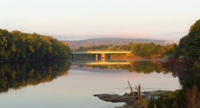View looking west upriver at I-93 bridge over Merrimack River in soft, pink morning light on wide, placid river in Concord, NH