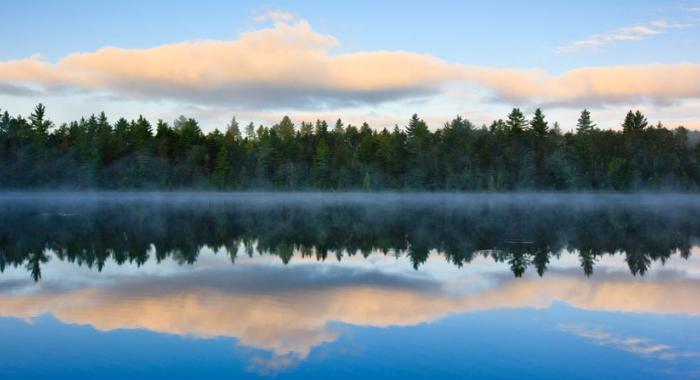 A forest and clouds are reflected in misty water below at Umbagog National Wildlife Refuge.