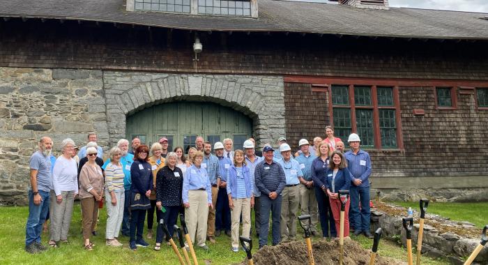 A large group poses behind a pile of dirt with ceremonial shovels, in front of the soon to be renovated Carriage Barn.