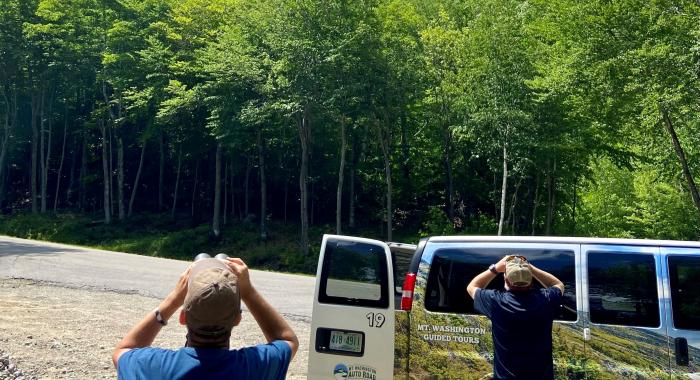 Two people use binoculars to look at the sky while the Mt Washington Auto Road van waits.