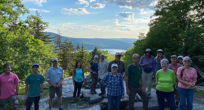 Colorful T-shirts of hikers in shadow at summit of Sunset HIll, view of Lake Sunapee in background