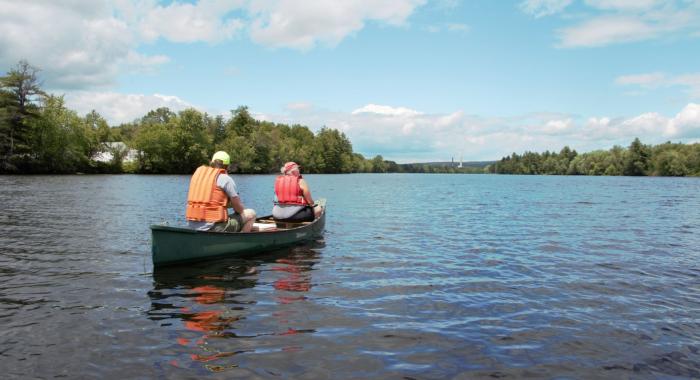 Two people in lifejackets in a canoe on the Merrimack River as seen from behind.