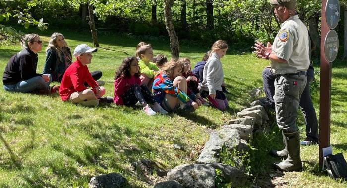 A group of school children sit on the lawn while a man speaks to them 