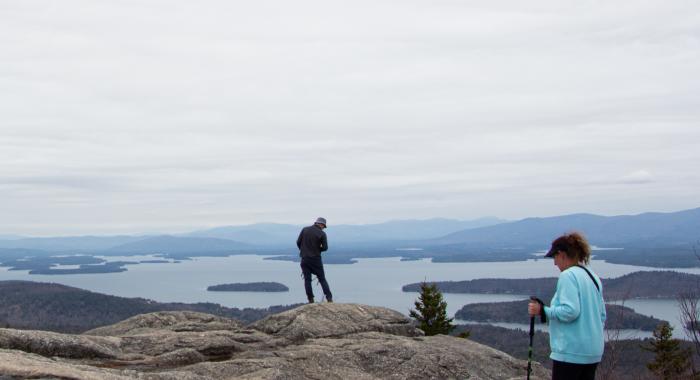 A woman in a blue shirt holding hiking poles hikes towards a man looking out on a mountain vista. 