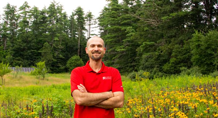 A man in a red shirt stands arms crossed in front of a large field 