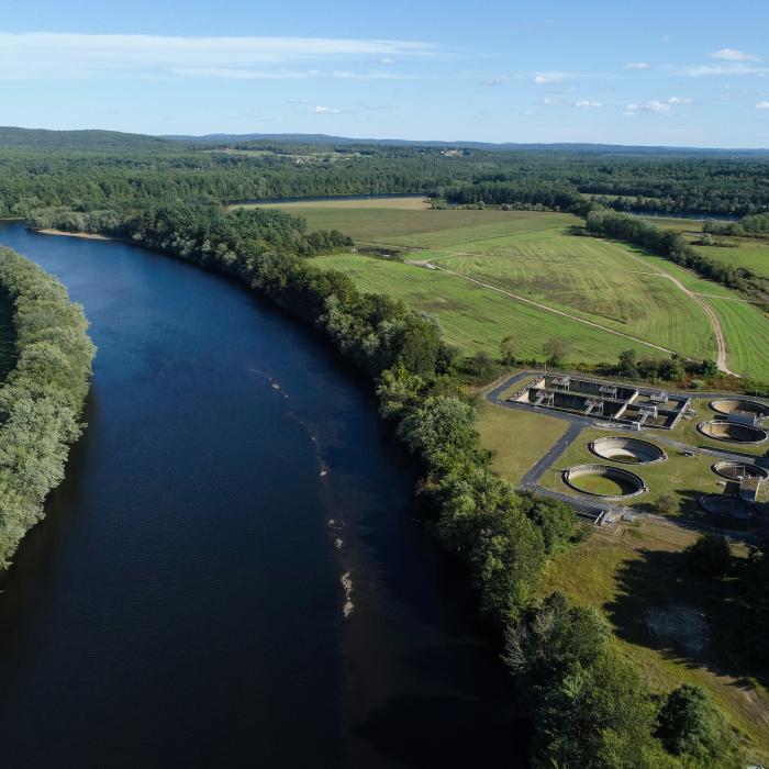 Blue river lined by trees on one side and gray buildings on the other.