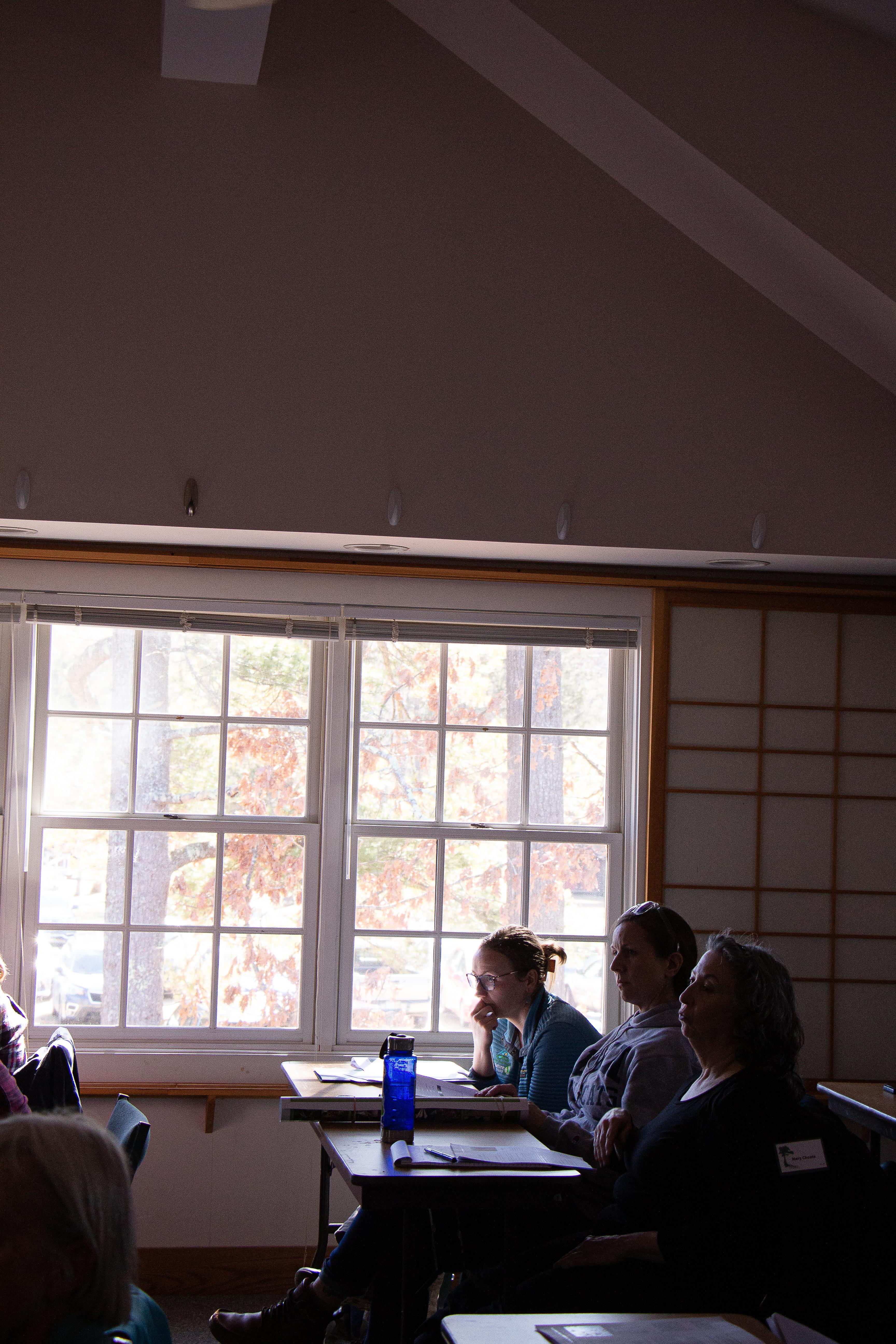 Three women sit at desks in front of a bright window.