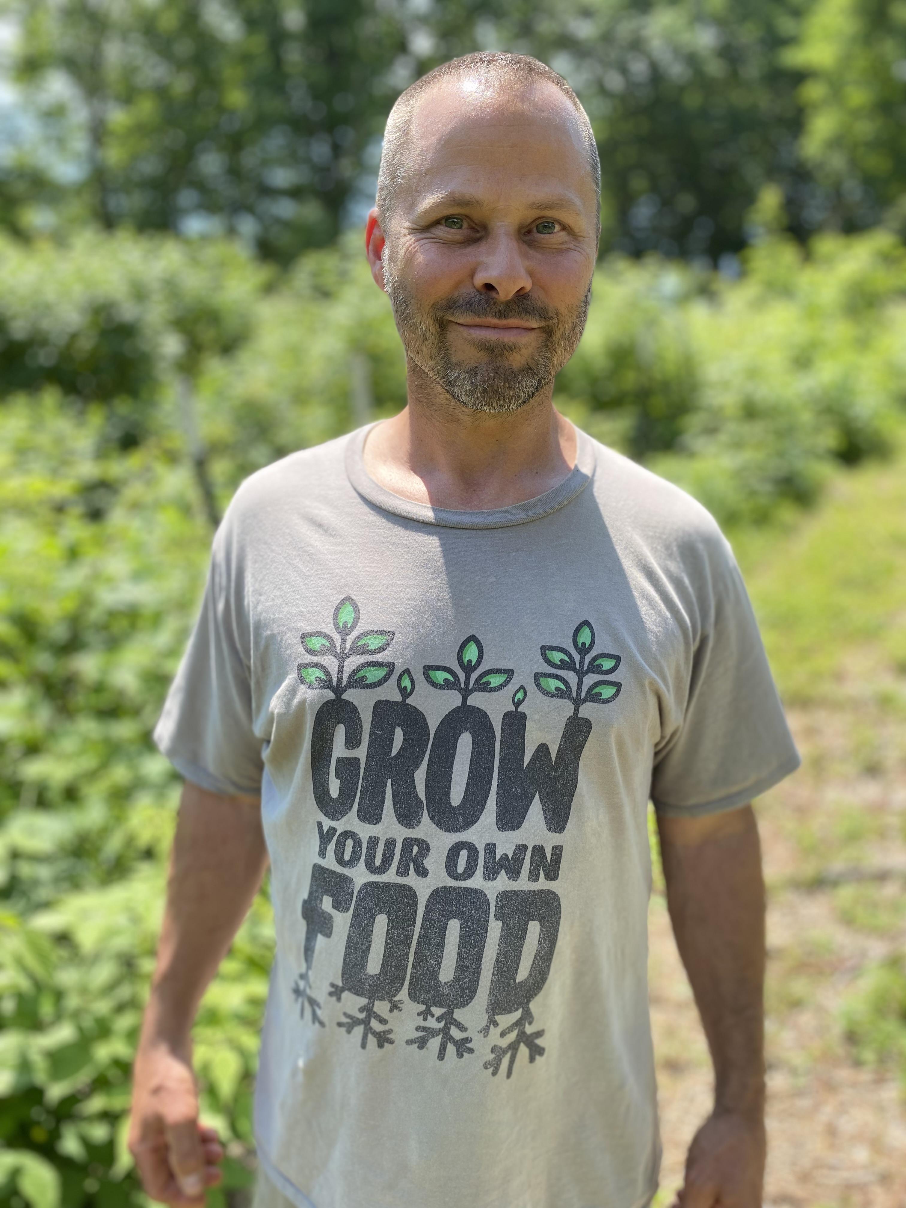 Man with beard stands in field wearing shirt that says "grow your own food"