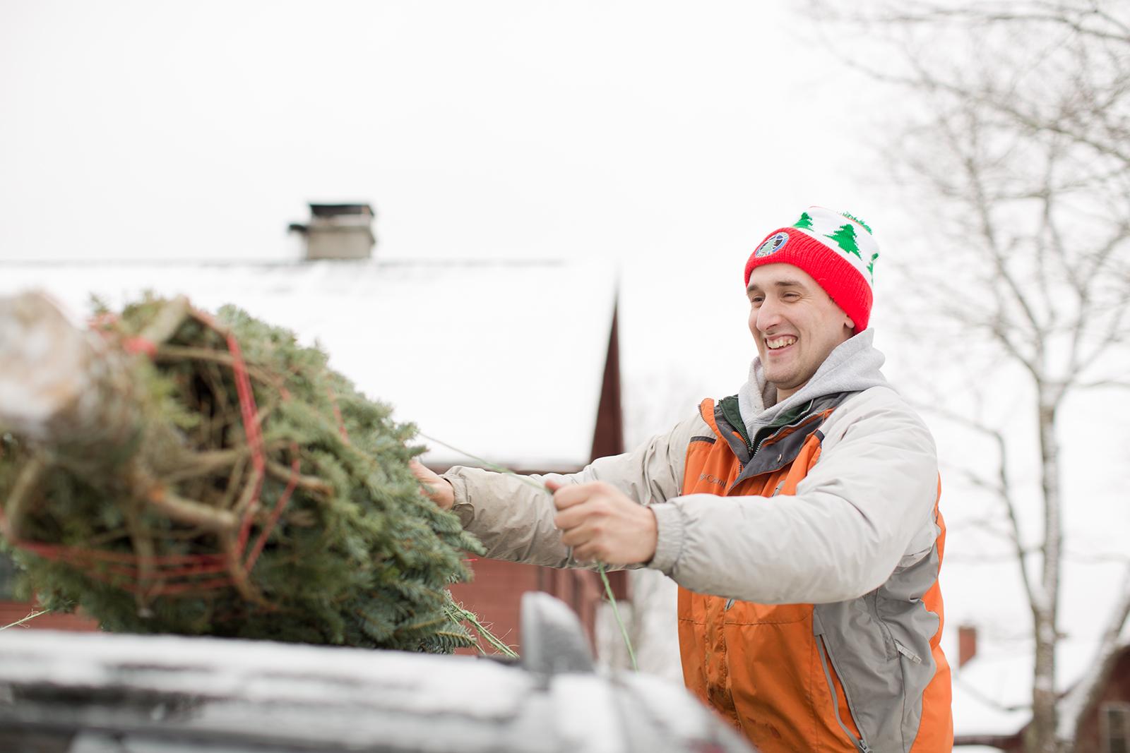 A snowy backdrop as a smiling person loads a Christmas tree on the baler