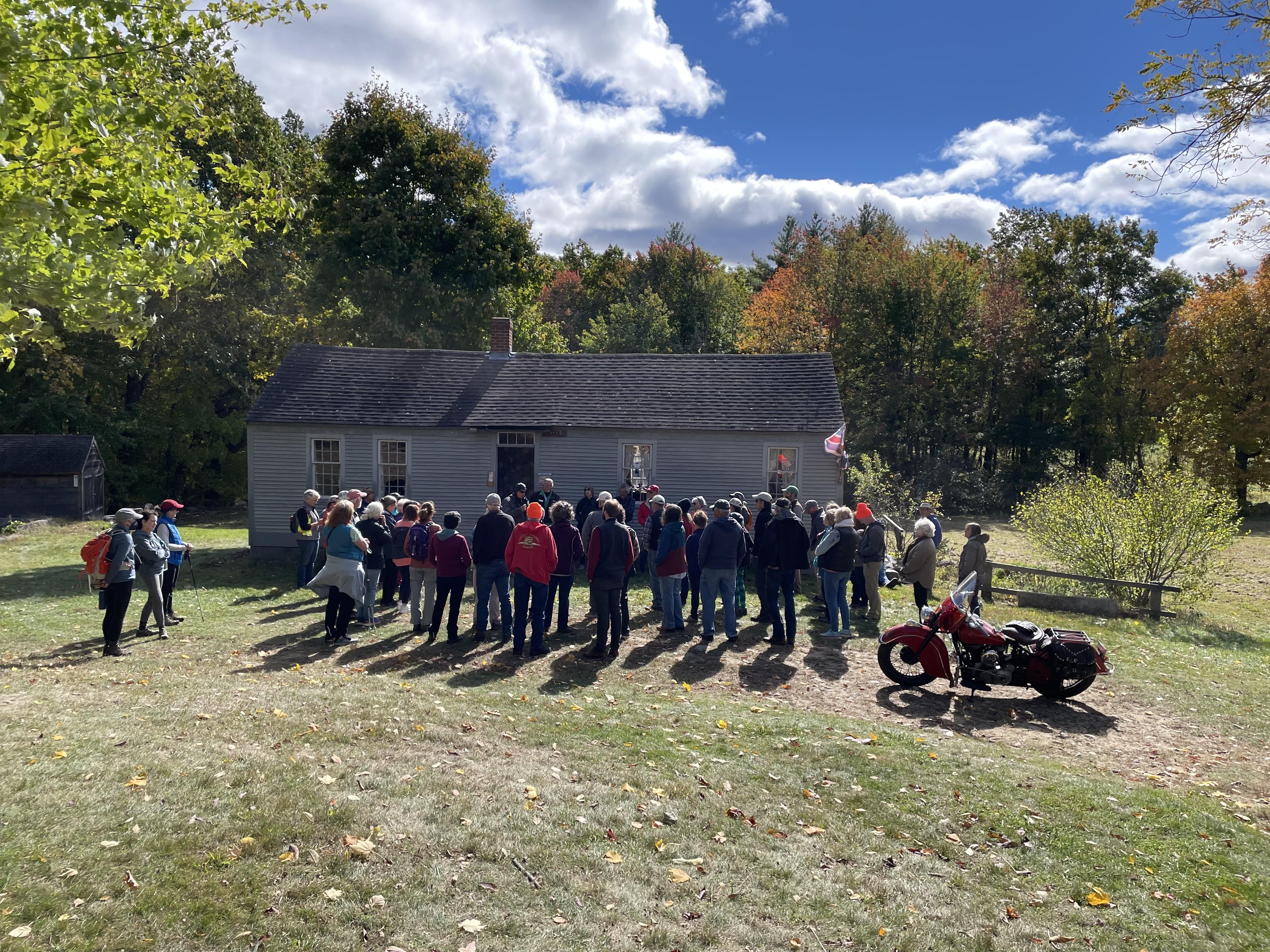 A group of people stand in front of a small grey house in a field on a sunny fall day.