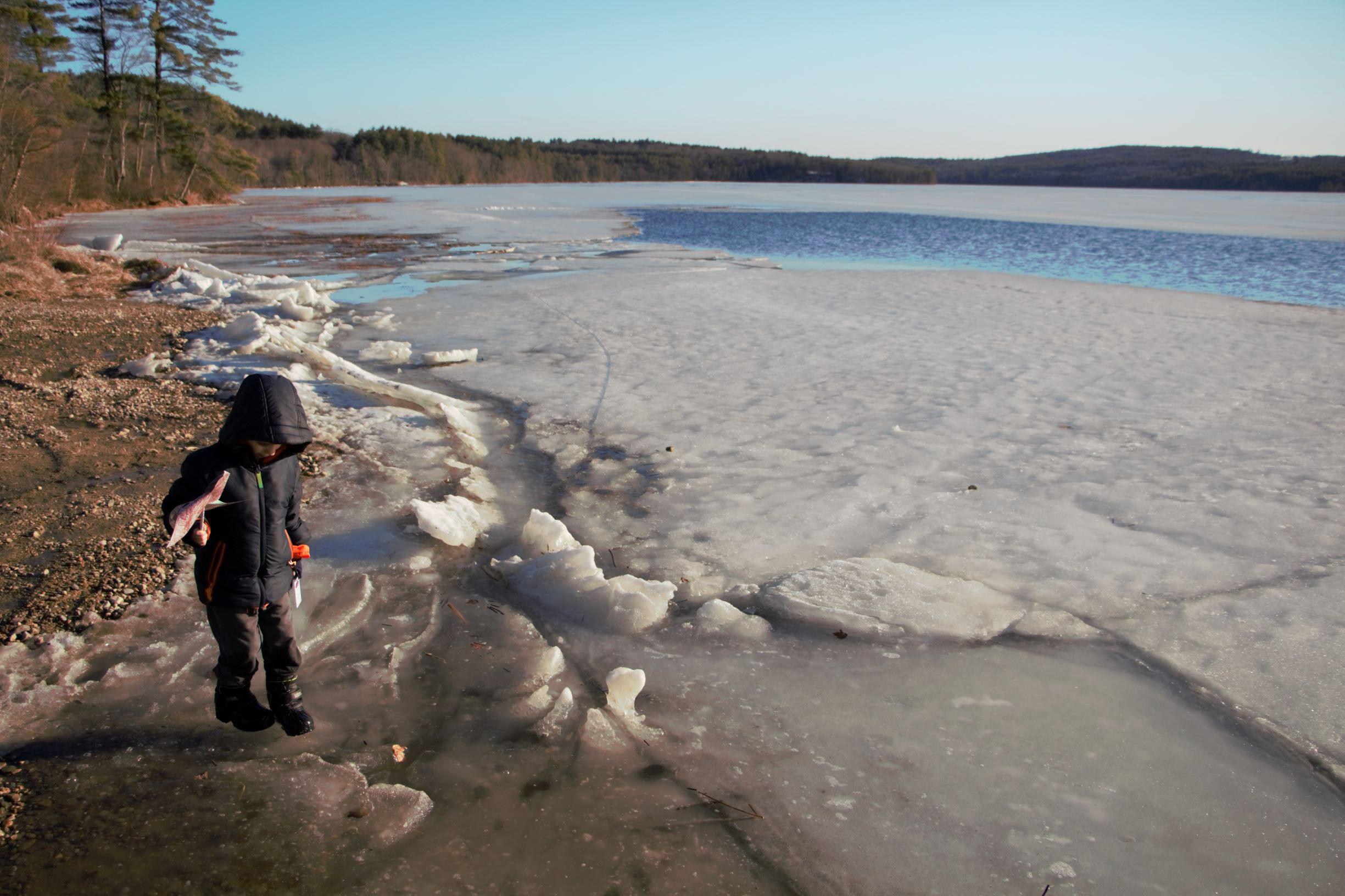 A boy in snow boots stands near an icy lake holding a pinwheel.