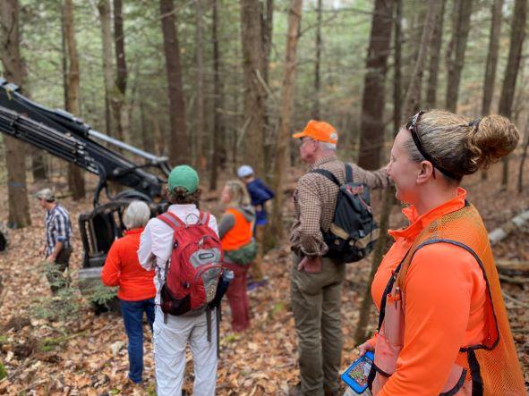 Blaze orange clad group view a logging machine in hemlock forest
