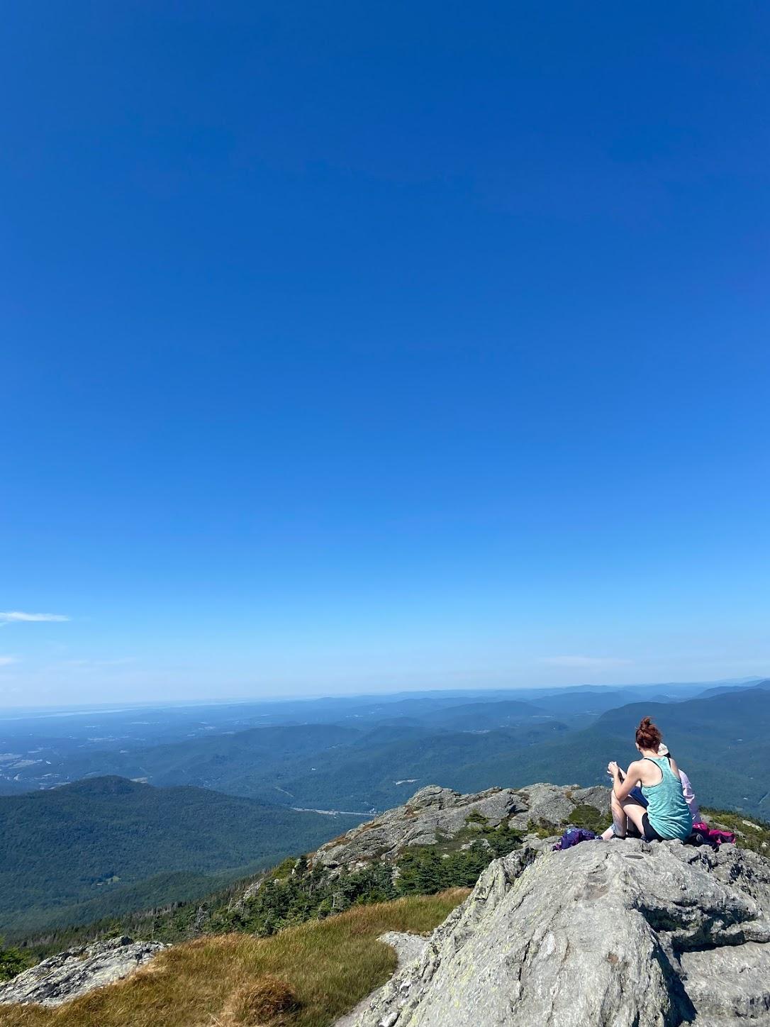 Woman sits on the edge of a large rock at the top of a mountain looking out to the view