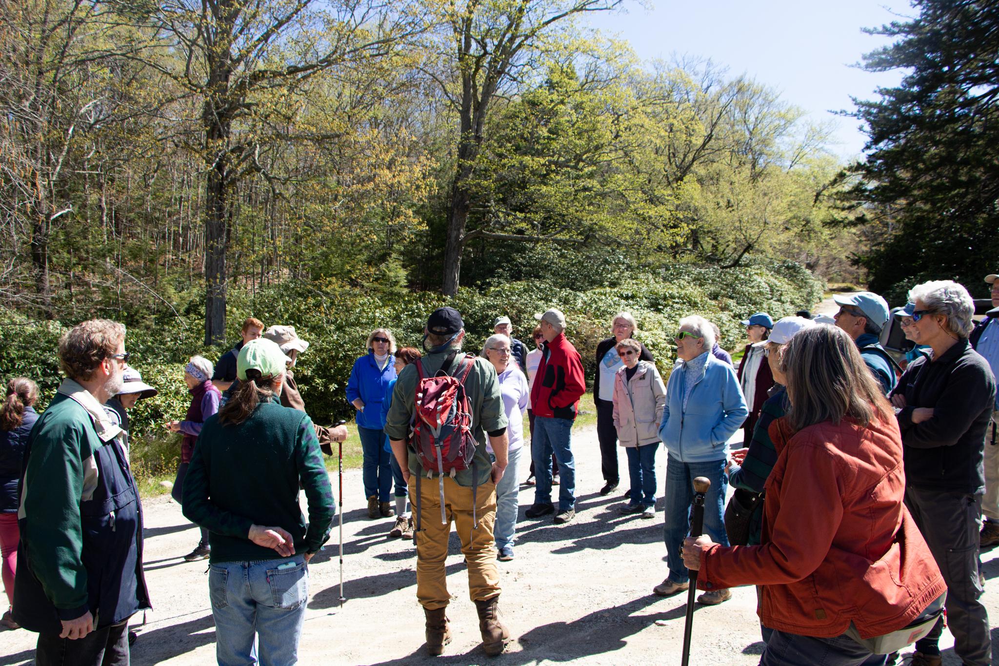 A man in a red backpack addresses a large crowd of people dressed for hiking.