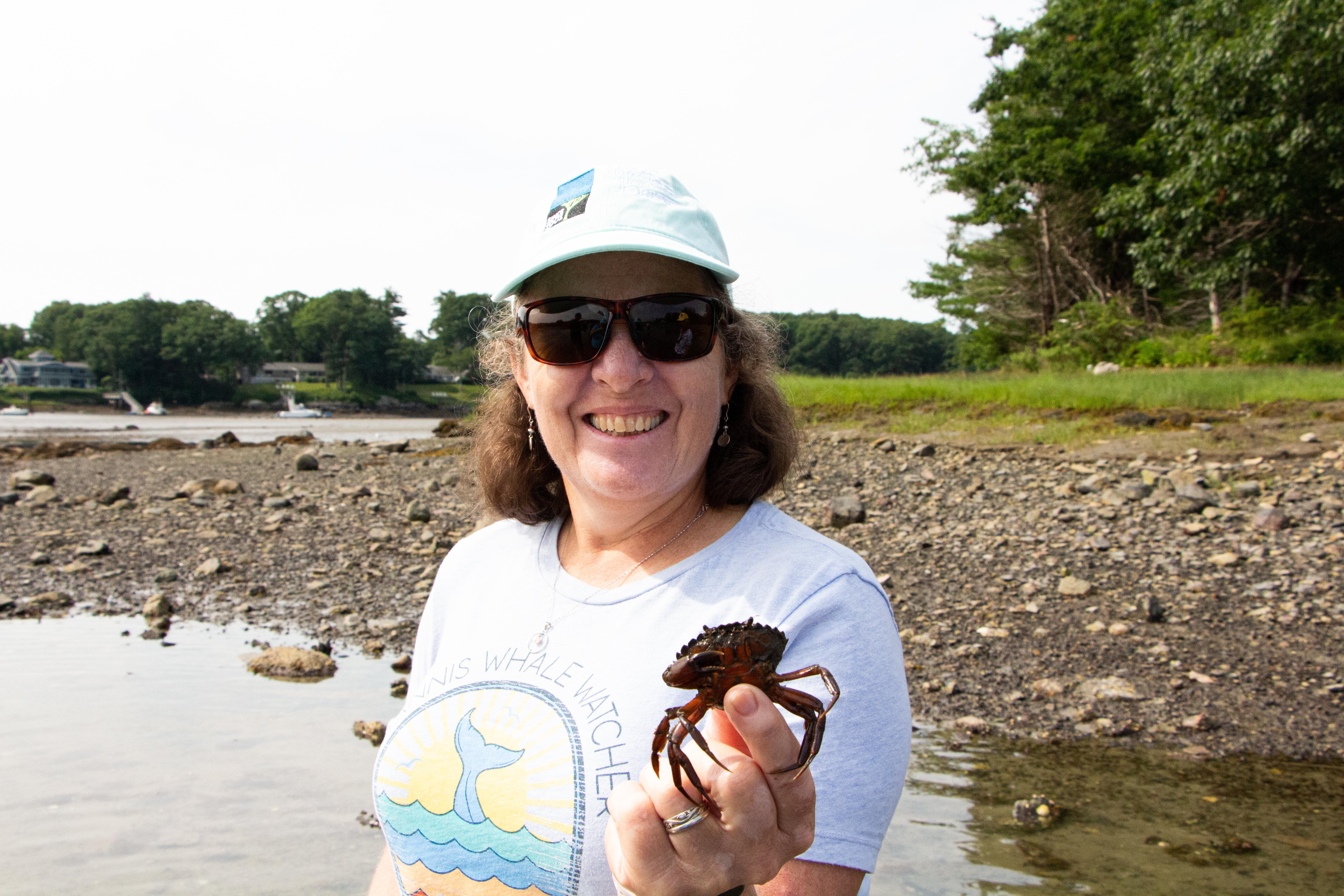 A woman wearing a baseball hat and sunglasses holds a crab out to the camera.