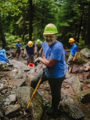 Trail volunteer poses with sledgehammer, eye protection, and a hardhat