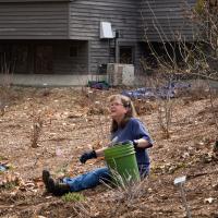 A woman sits in a garden, holding a green bucket. 