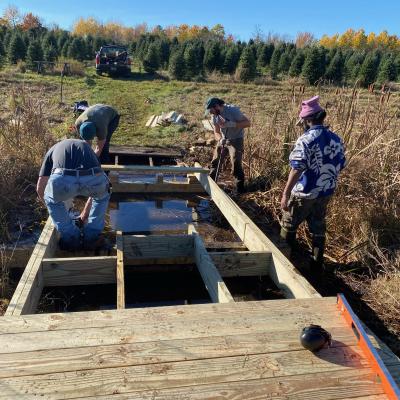 Four people work on a large footbridge in a Christmas tree farm field.