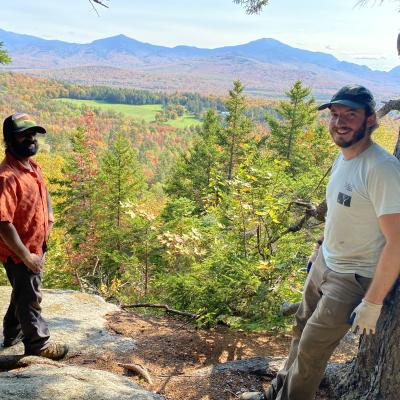 Two men stand on a rock overlooking a mountain view.
