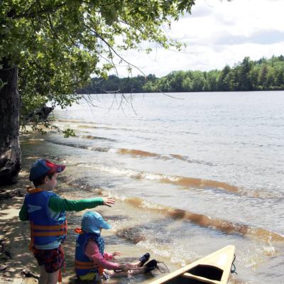 Two boys sit with their feet in the water of the Merrimack River near a forested shore.