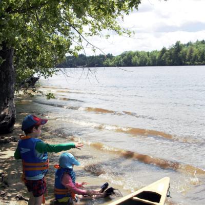 Two boys sit with their feet in the water of the Merrimack River near a forested shore.