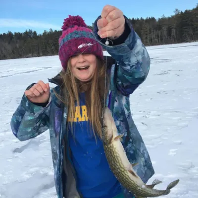 A young girl in winter clothes holds up a long fish on a fishing line