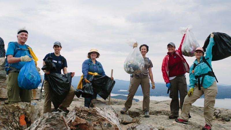 A group of individuals holding full trash bags at the summit of a mountain on a grey sky day.