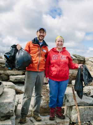 Two individuals holding up full trash bags on a rocky area with a blue sky background.