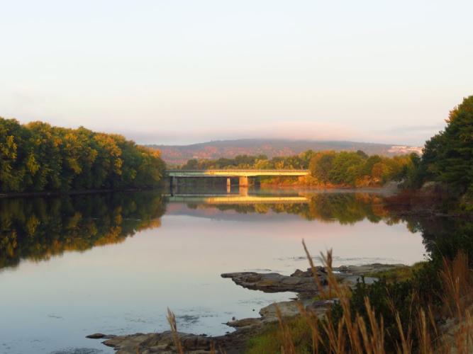 View looking west upriver at I-93 bridge over Merrimack River in soft, pink morning light on wide, placid river in Concord, NH