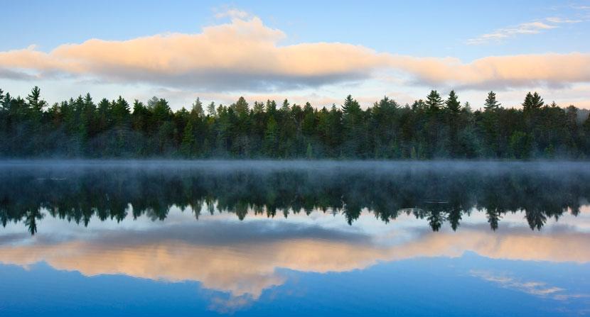 A forest and clouds are reflected in misty water below at Umbagog National Wildlife Refuge.