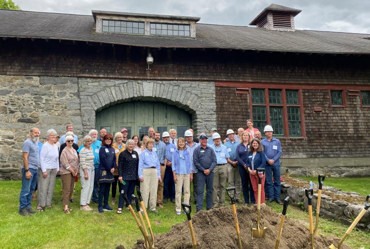A large group poses behind a pile of dirt with ceremonial shovels, in front of the soon to be renovated Carriage Barn.