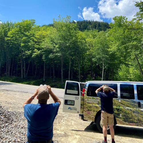Two people use binoculars to look at the sky while the Mt Washington Auto Road van waits.