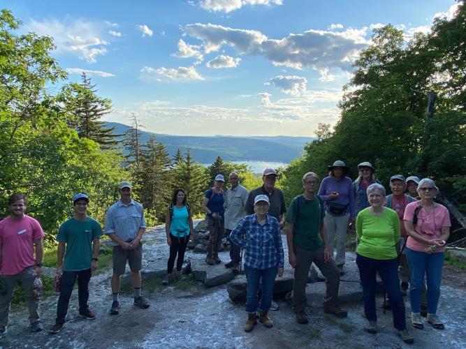 Colorful T-shirts of hikers in shadow at summit of Sunset HIll, view of Lake Sunapee in background
