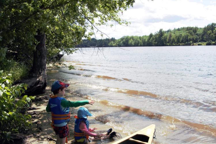 Two boys sit with their feet in the water of the Merrimack River near a forested shore.