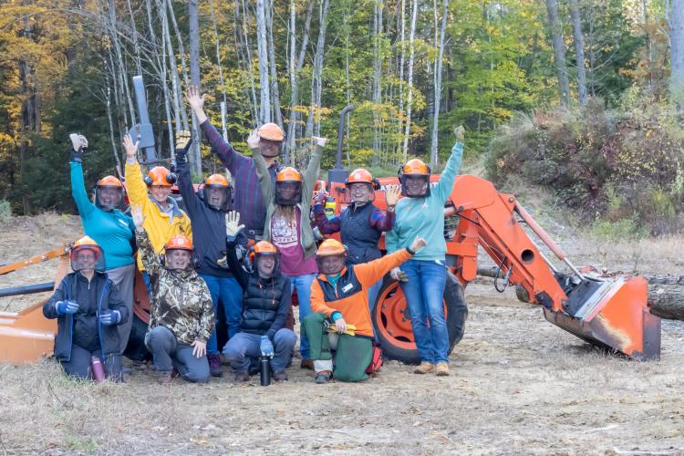 A group of female identifying people gather together for a photo, clad in orange hardhats, in front of a tractor during a Women in the Woods workshop.